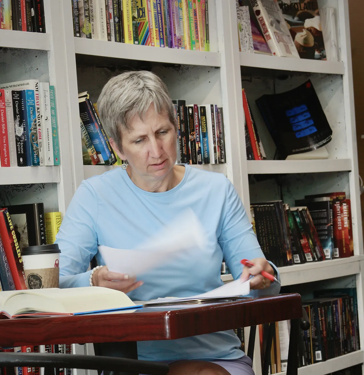 woman in blue shirt sitting at a table in front of a wall of books editing document by hand with red pen