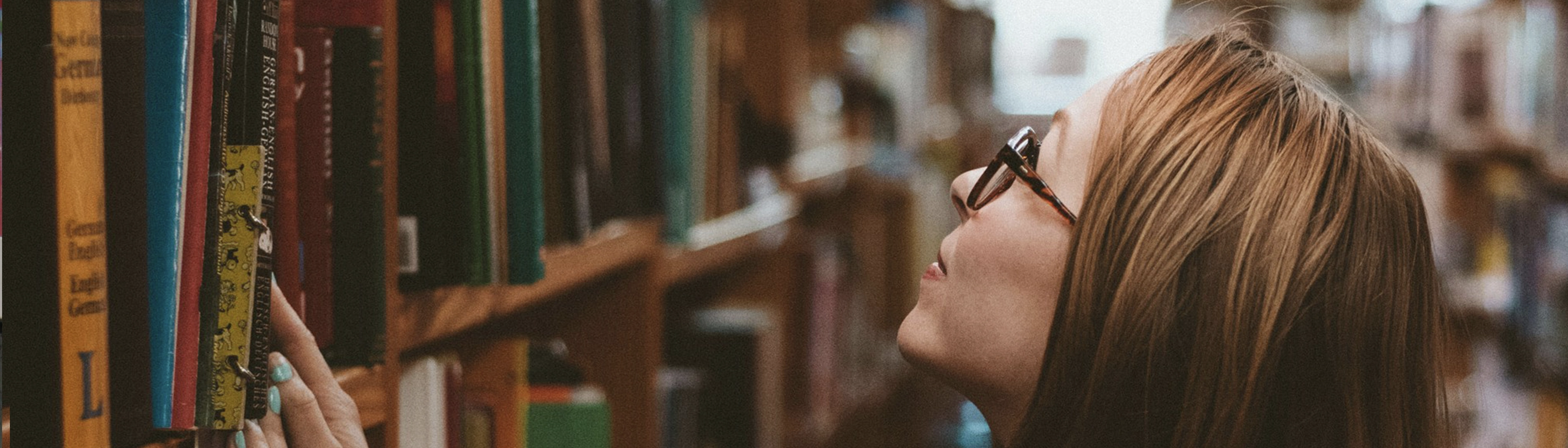 A woman with brown hair and glasses, in a bookstore. She is standing in front of a shelf of books, looking up at a shelf.