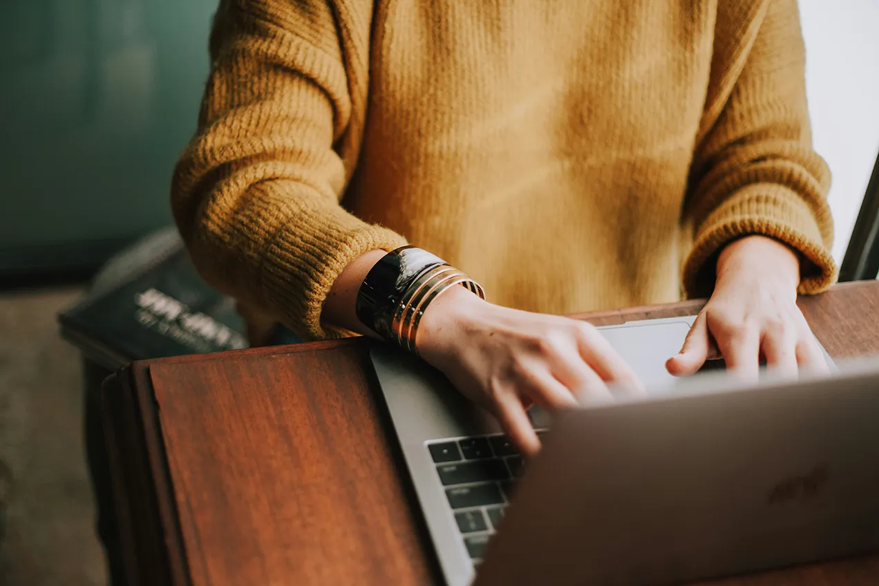 woman typing on a laptop wearing a mustard colored sweater and silver bracelets