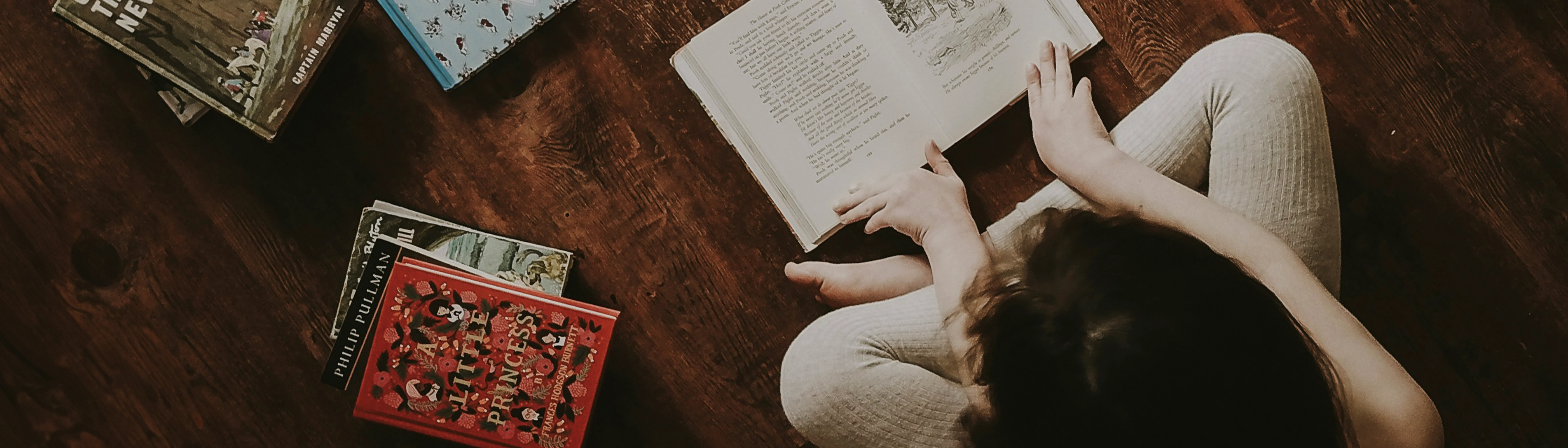 A person sitting on wooden floor reading a book, surrounded by book stacks.